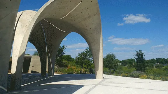 Curved concrete pavilion at Confluence Park, captured from low angle under bright blue sky with scattered clouds.